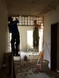 Zwei Bauarbeiter stehen auf Leitern und arbeiten an einer Betondecke, Funken fliegen, Baustelle mit Trümmern im Hintergrund.
