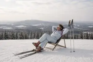 Mann in Skikleidung entspannt auf einem Liegestuhl im Schnee mit verschneiten Bergen und Tannenbäumen im Hintergrund.