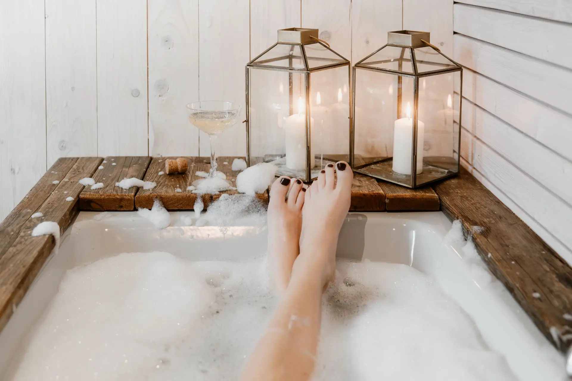 Füße einer Person in einer Badewanne mit Schaum, umgeben von Kerzen und einem Glas Champagner auf einer Holzablage.