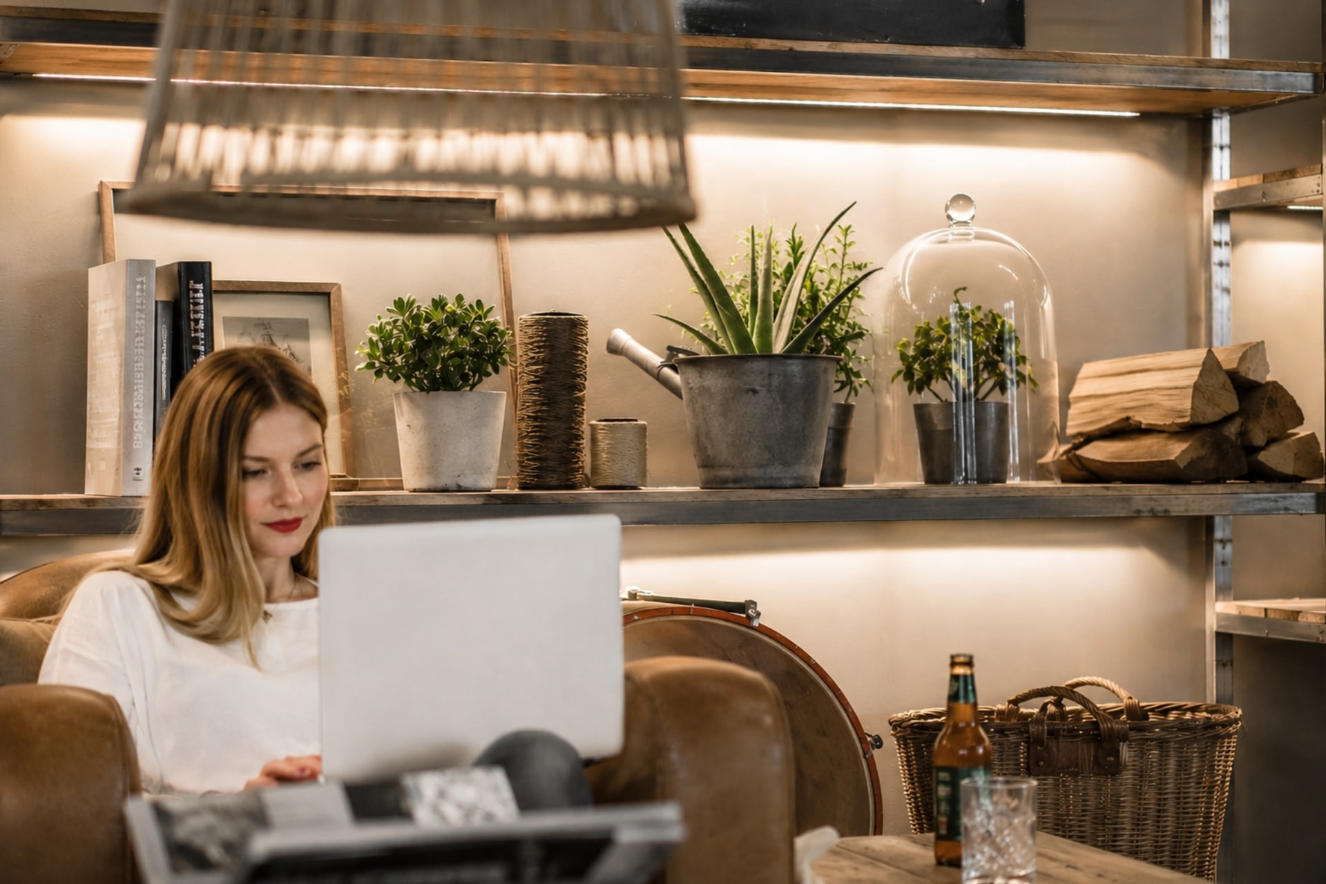 Frau mit langer blonden Haaren sitzt auf einem Sessel, arbeitet am Laptop, umgeben von Pflanzen und Holz.