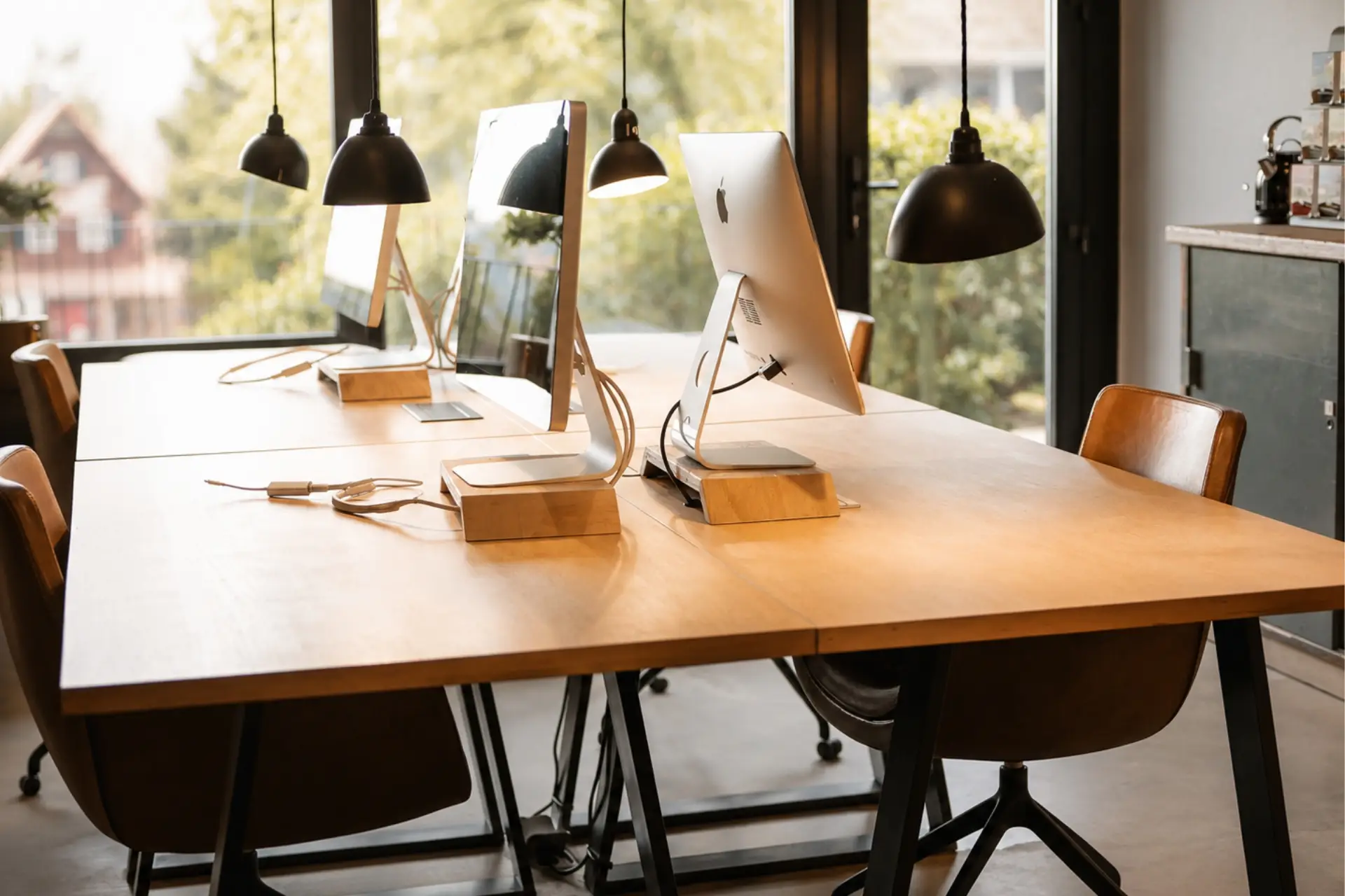 Büro mit hellen Holztischen, zwei Apple-Computern, modernen Lampen und Blick auf einen Garten. Kunststoffkabel sichtbar.