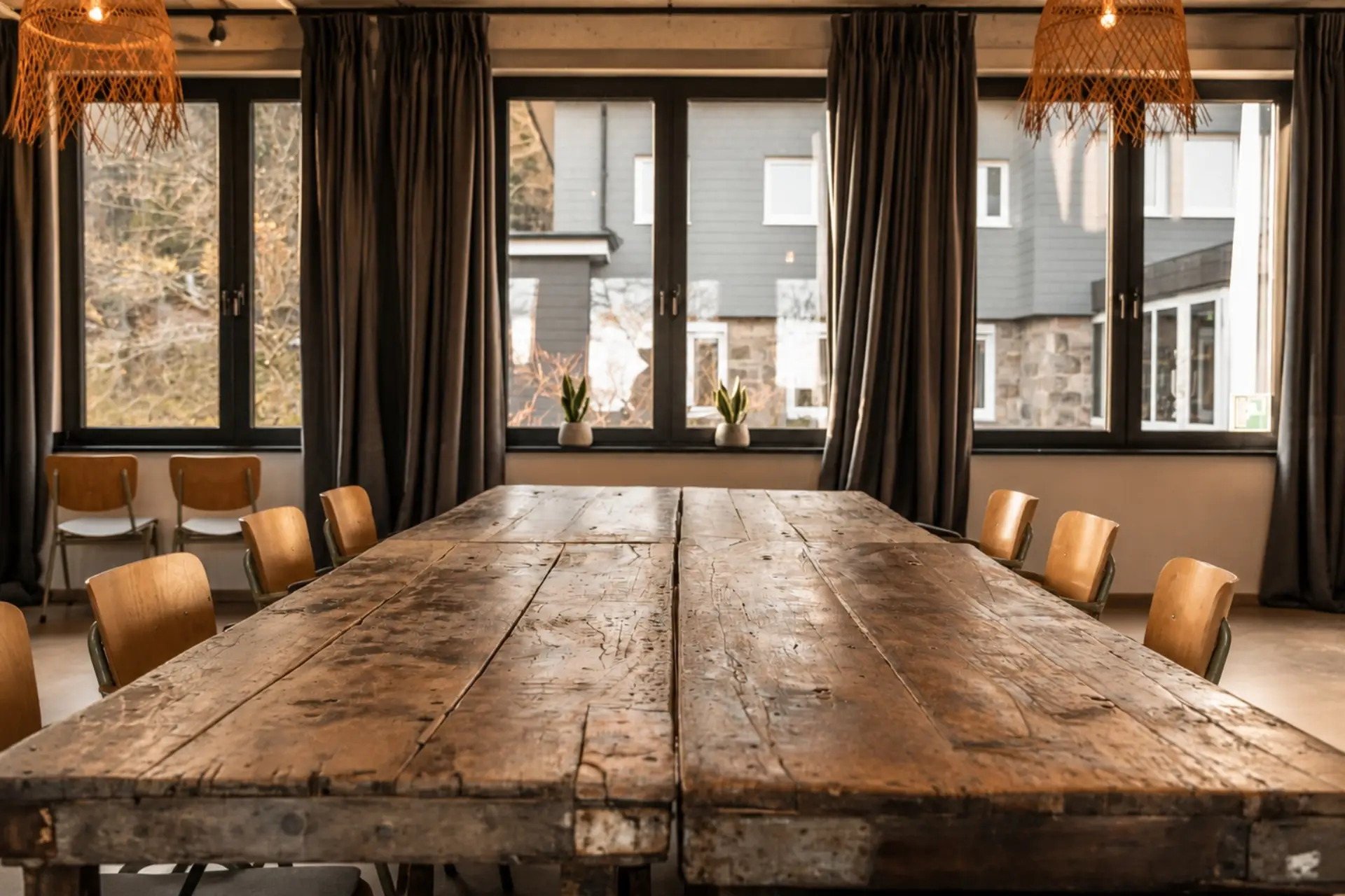 Gemütlicher Besprechungsraum mit einem großen Holztisch und Stühlen, Blick auf die Fenster mit Pflanzen.