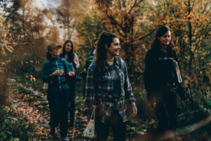 Vier Frauen wandern fröhlich einen schmalen Pfad durch einen herbstlichen Wald, umgeben von buntem Laub.